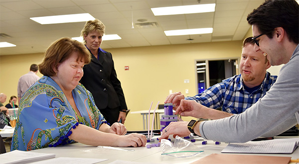 People working together to build the tallest tower at a desk while the instructor looks at the team's results. 