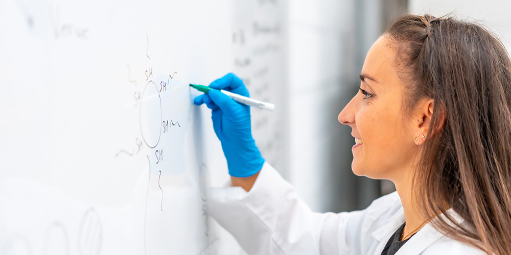 Woman in lab coat writing chemical equations on a white board.